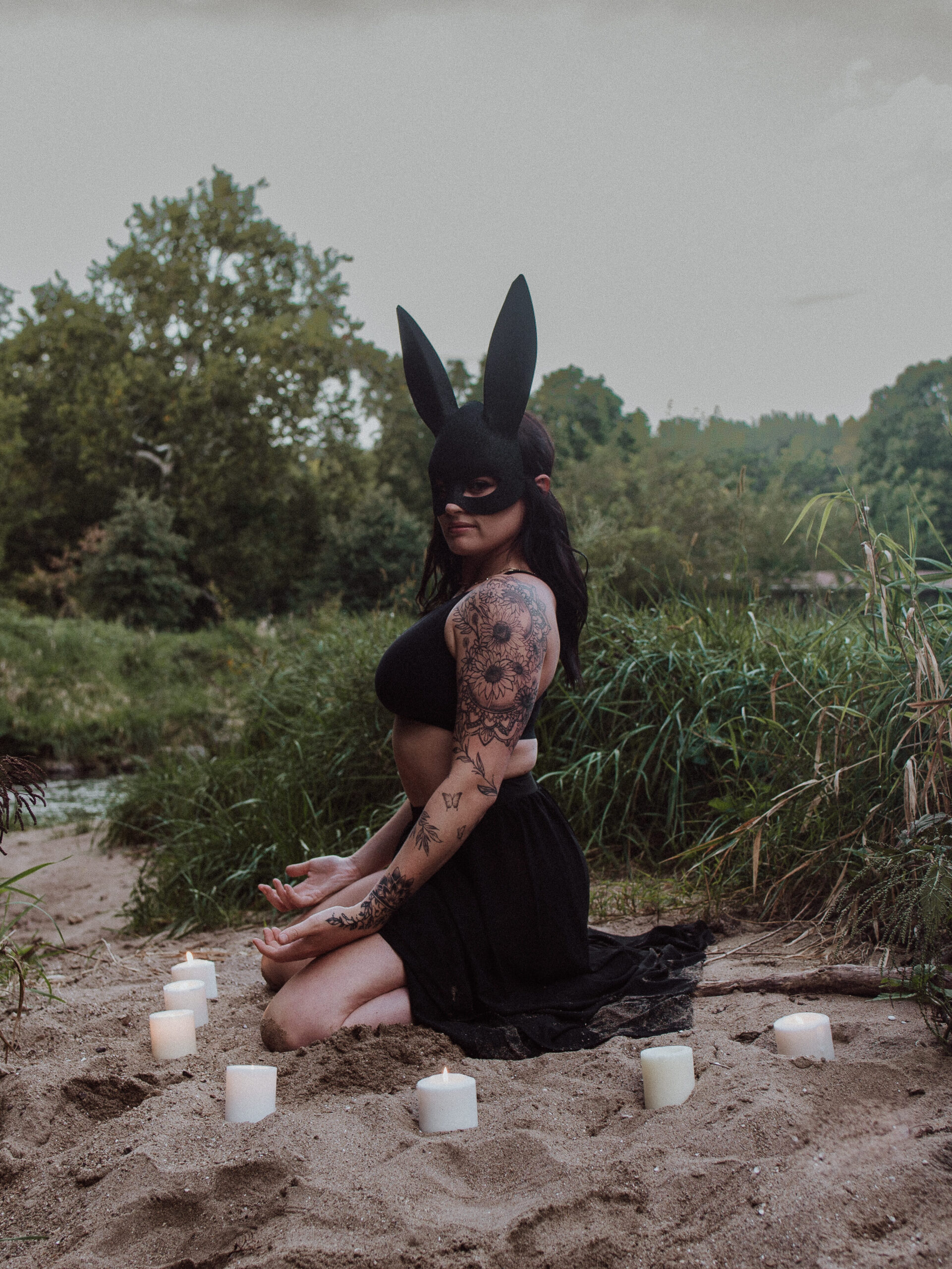 Woman sitting on the sand near the water with candles surrounding her in a circle wearing a half black bunny mask